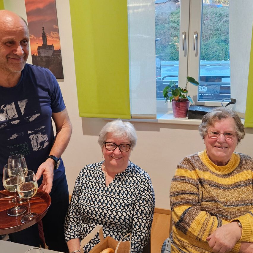 Three people are smiling in a room with yellow blinds and a window showing trees. A man holds wine glasses, a woman holds a bag, and another woman sits on a bench.