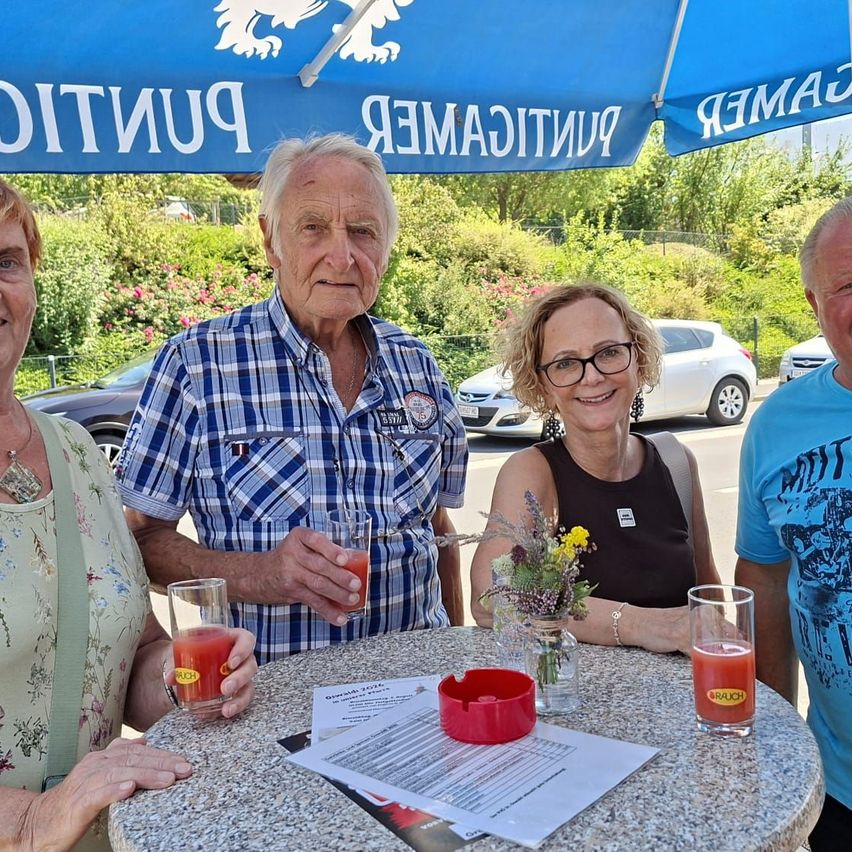 Vier Personen sitzen um einen Tisch unter einem blauen Schirm, lächeln und halten Getränke. Eine Frau links trägt ein Blumenhemd, während der Mann neben ihr ein blaues kariertes Hemd trägt. Eine Frau in der Mitte trägt eine Brille und ein schwarzes Tanktop. Der Mann rechts trägt ein blaues Shirt mit einem grafischen Design. Hinter ihnen sind Autos geparkt und ein Zaun mit Grünpflanzen.