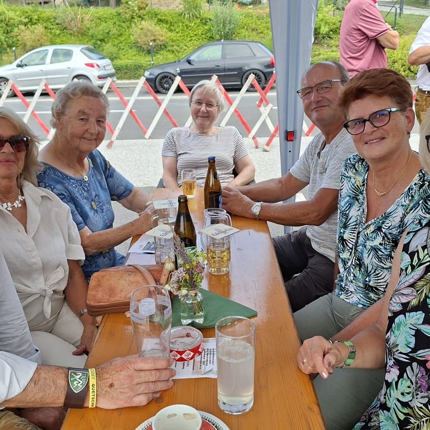 Eine Gruppe älterer Menschen sitzt an einem Tisch mit Getränken und Essen. Sie lächeln und scheinen ihre Mahlzeit zu genießen. Im Hintergrund sind Autos geparkt.