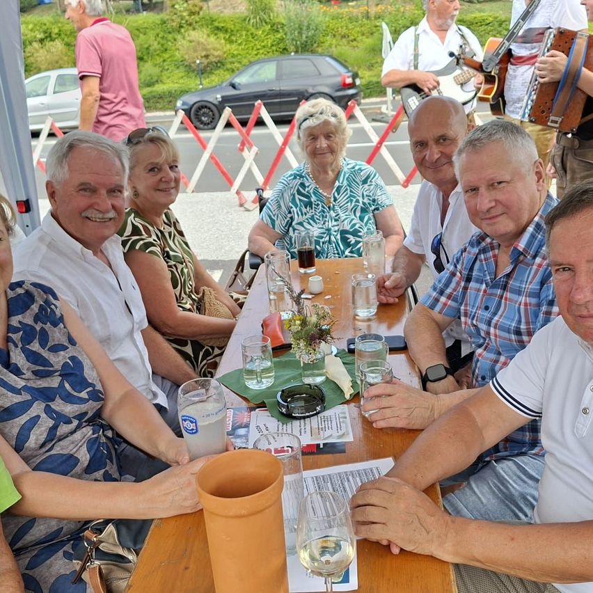 Eine Gruppe älterer Erwachsener sitzt um einen Tisch herum, lächelt und posiert für ein Foto. Der Tisch ist mit Gläsern, einer Blumenarrangement und verschiedenen Gegenständen gedeckt. Hinter ihnen spielt eine Person Akkordeon. Im Hintergrund sind geparkte Autos und ein Grasbereich zu sehen.