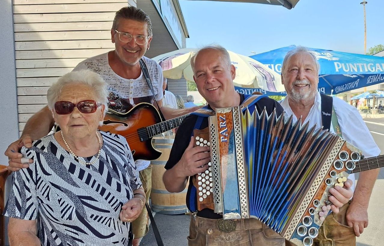 Vier Personen lächeln und posieren für ein Foto. Ein Mann spielt Gitarre, ein anderer Akkordeon, und zwei weitere stehen neben ihnen. Sie befinden sich in einer Außenumgebung mit Schirmen und Fässern im Hintergrund.