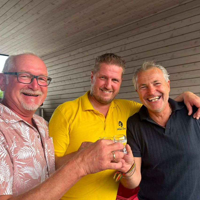 Drei Männer lächeln und posieren für ein Foto, einer hält ein Glas, ein anderer hat seinen Arm um die Schultern des Mannes auf der rechten Seite gelegt.