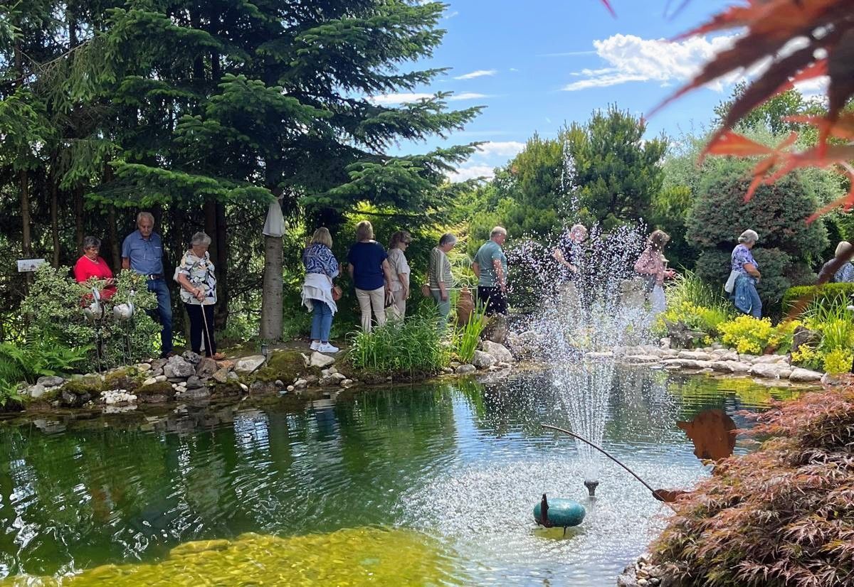 Eine Gruppe von Menschen versammelt sich um einen Teich und bewundert den Springbrunnen in der Mitte. Bäume und Pflanzen umgeben den Bereich.