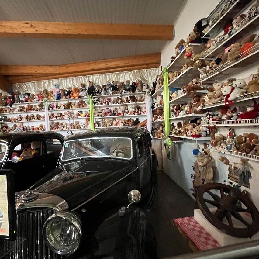 Ein Raum mit Holzbalken zeigt einen schwarzen Oldtimer in der Mitte geparkt, mit Regalen voller Kuscheltiere an den Wänden.