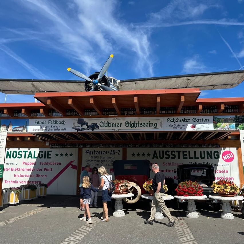 Eine Outdoor-Touristenattraktion mit einem Oldtimer-Flugzeug. Leute stehen vor dem Eingang, und es gibt Blumendisplays und Oldtimer-Autos zu sehen.
