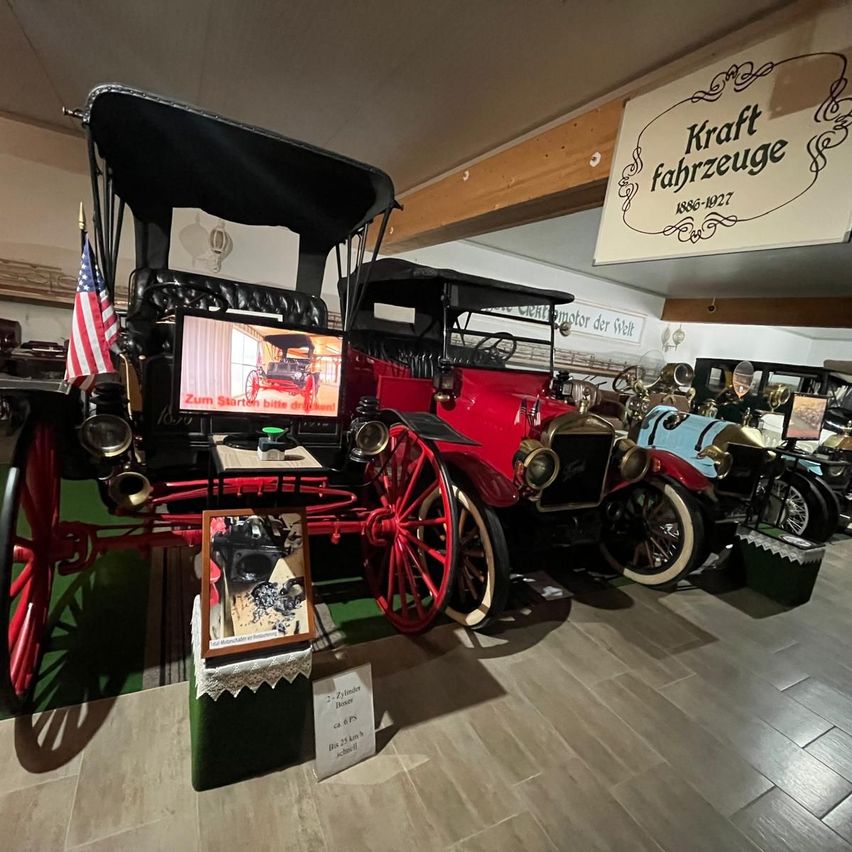 Ein rotes Oldtimerauto wird in einem Museum ausgestellt. Das Auto steht auf einer grünen Plattform mit einer amerikanischen Flagge. Über dem Auto befindet sich ein Schild mit der Aufschrift 'Kraftfahrzeuge 1886-1927'.
