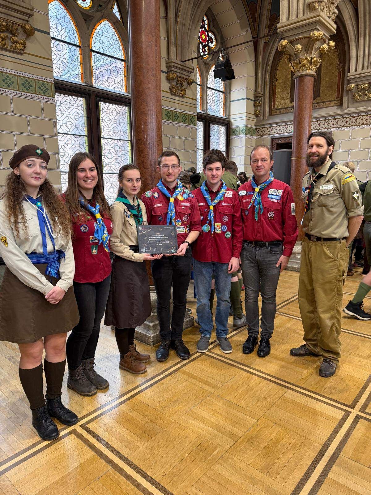 A group of scouts, including two women and four men, pose with a plaque in a formal setting. They are wearing uniforms and standing on a wooden floor.