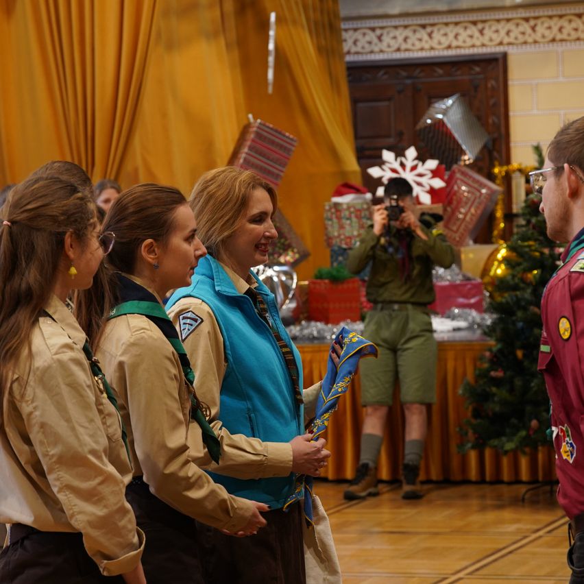 A group of women in scout uniforms stands next to a man. A photographer captures the moment in the background, with decorations and gifts nearby.