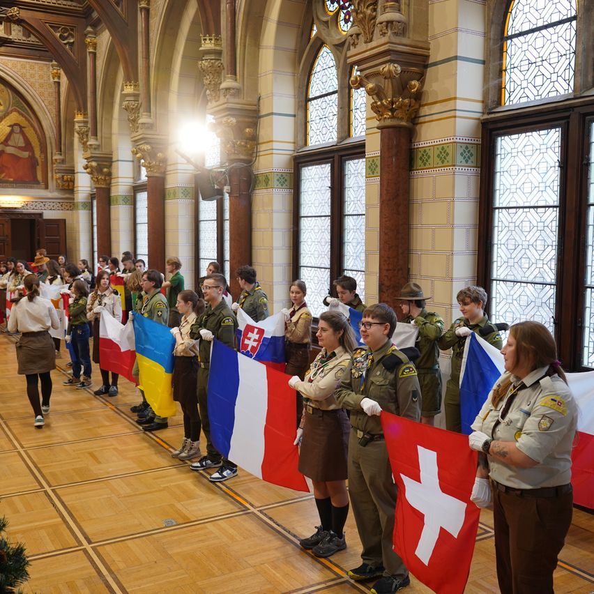 A group of scouts stands in a row, holding flags from various countries. They wear uniforms and are inside a room with large arched windows.