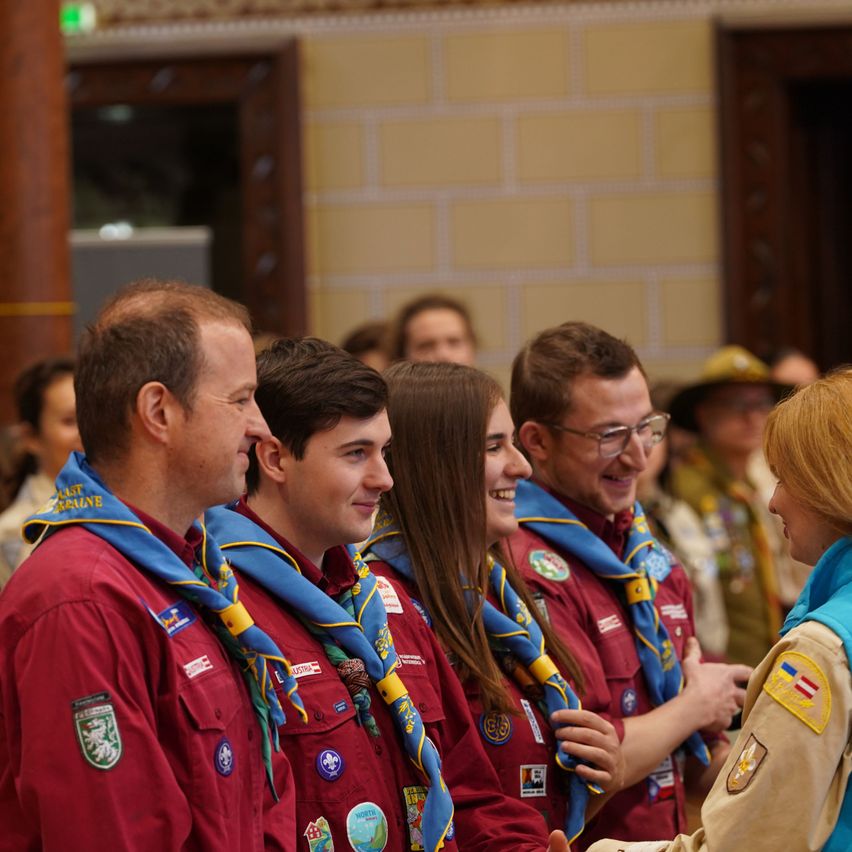 A group of smiling scouts, both male and female, wearing red uniforms with badges, stand in a line inside a building. They appear to be conversing with a woman in a beige uniform.