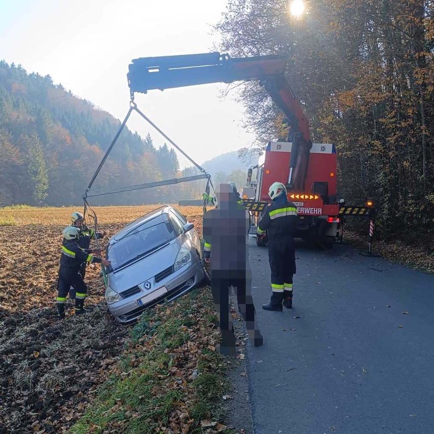Feuerwehrleute benutzen einen Kran, um ein beschädigtes Fahrzeug von der Straße zu heben. Drei Personen in Schutzkleidung helfen. Ein Feuerwehrwagen ist in der Nähe geparkt.