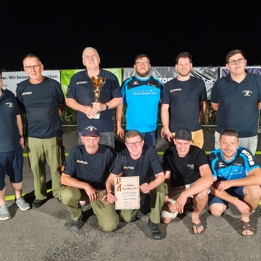A group of men in uniforms and casual wear pose for a photo with a trophy at an outdoor event.