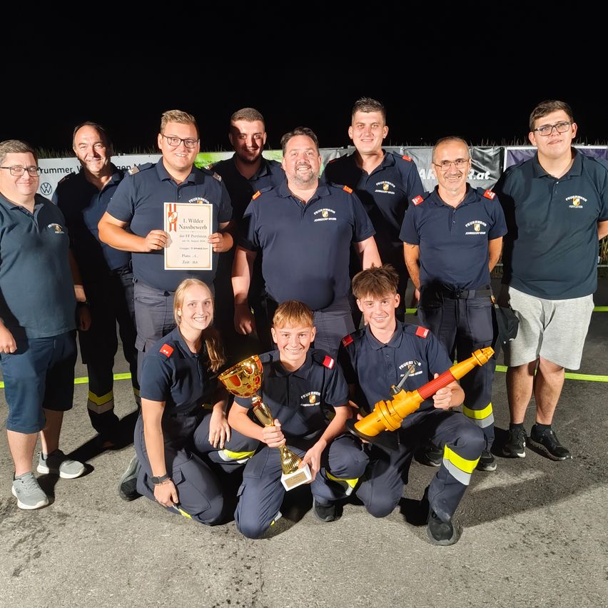 A group of firefighters pose for a photo at night, with some holding trophies and certificates. They are all wearing uniforms and smiling.