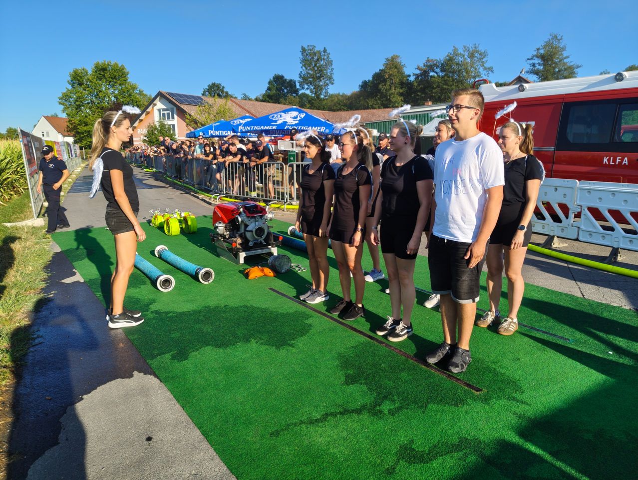 A group of people dressed in black outfits stand on a green carpet, possibly participating in an event. A man in a white shirt stands in front of them. Behind them, there is a crowd of people watching.