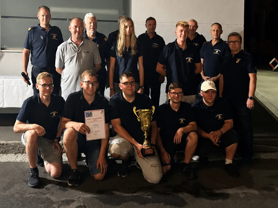 Eine Gruppe von Menschen in Uniform, einige mit Mützen, posiert für ein Foto mit einem Pokal und einem Zertifikat. Sie lächeln und stehen vor einer weißen Wand.