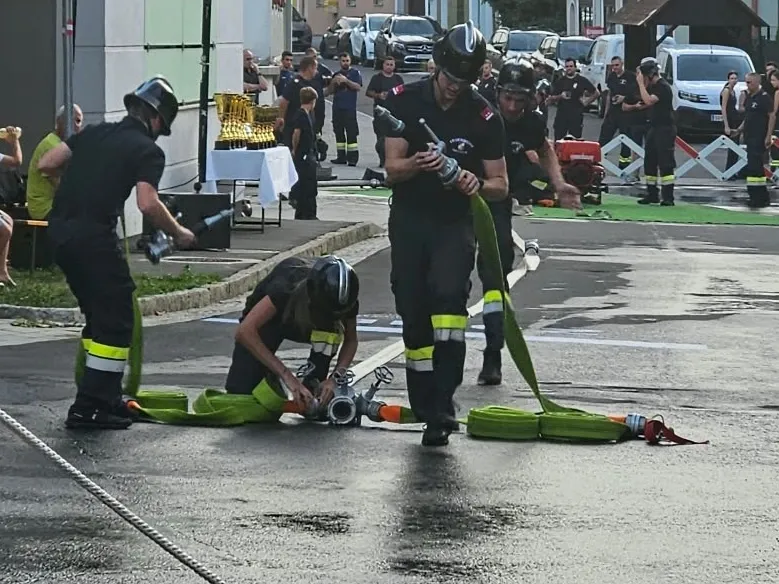 Feuerwehrleute üben auf einer nassen Straße, eine Person kniet und eine andere hält eine Feuerwehrspritze. Zuschauer beobachten vom Bürgersteig aus.