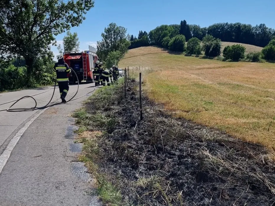 Feuerwehrleute sind am Straßenrand und löschen ein Feuer in einem Grasfeld. Ein roter Feuerwehrwagen ist in der Nähe geparkt.
