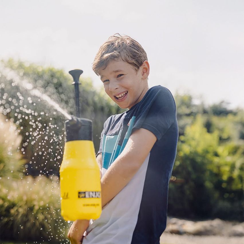 Ein Junge hält einen gelben Wassersprüher und sprüht Wasser, während er in einem Garten mit grünen Pflanzen lächelt.
