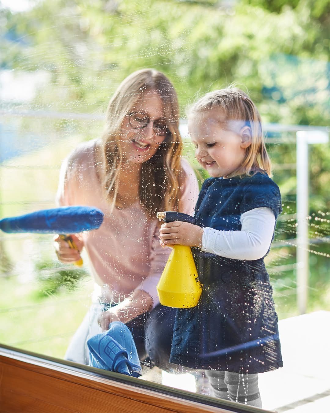 Eine Mutter und Tochter putzen ein Fenster mit Sprühflaschen und Tüchern, beide lächeln und genießen die Aktivität.