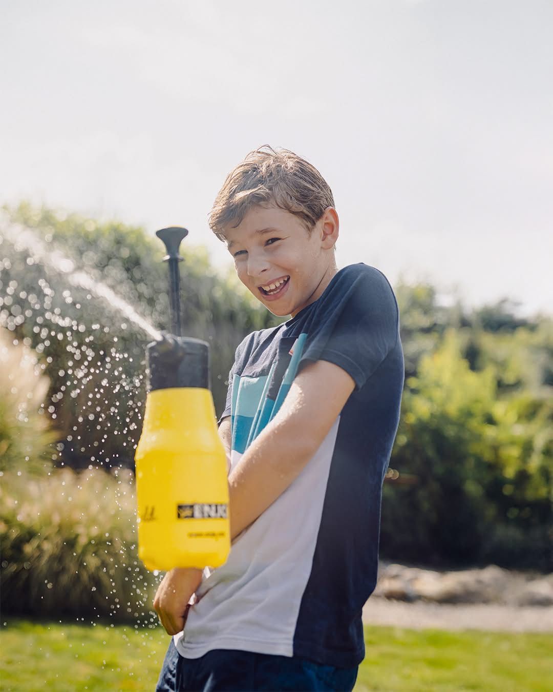 Ein Junge sprüht Wasser aus einem gelben und schwarzen Gießkannen in einem Garten mit grünen Pflanzen.