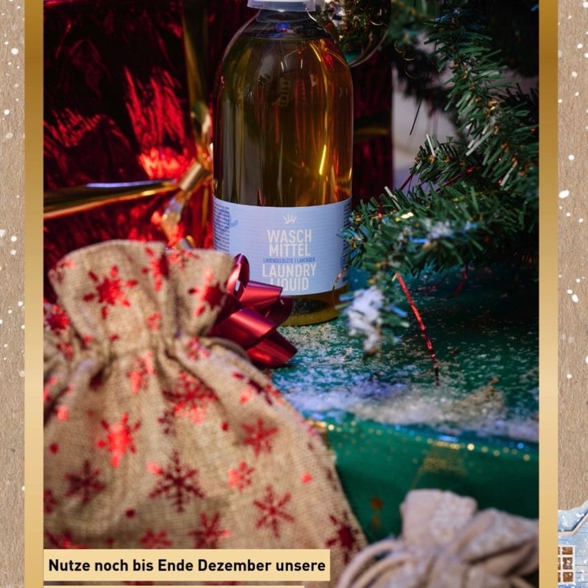 Eine Flasche Lavendel-Waschmittel ist in ein rotes Band gewickelt und auf einer grünen Oberfläche mit einem Weihnachtsbaum im Hintergrund platziert. Der Baum ist mit roten und goldenen Ornamenten geschmückt.