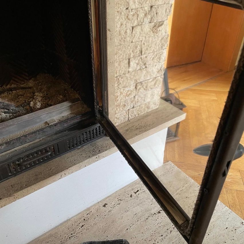 Ein Kamin mit offener Tür, der eine leere Feuerstelle mit Asche und Trümmern im Inneren zeigt. Der Kamin ist an einer Steinmauer befestigt.