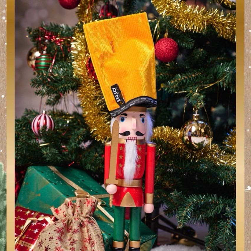 Ein Nussknacker-Soldat mit einem gelben Hut steht vor einem geschmückten Weihnachtsbaum. Er hält ein Schwert in der Hand und ist von verpackten Geschenken umgeben.