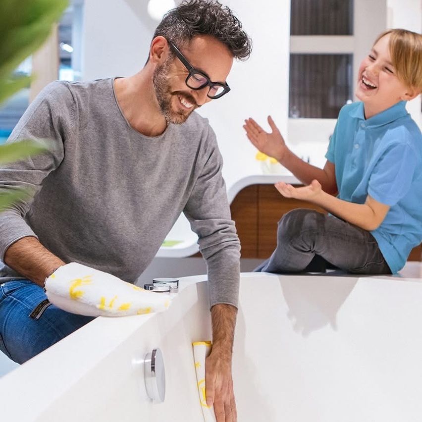 Ein Mann mit Brille reinigt eine Badewanne, während ein Junge in Blau daneben sitzt und lacht.