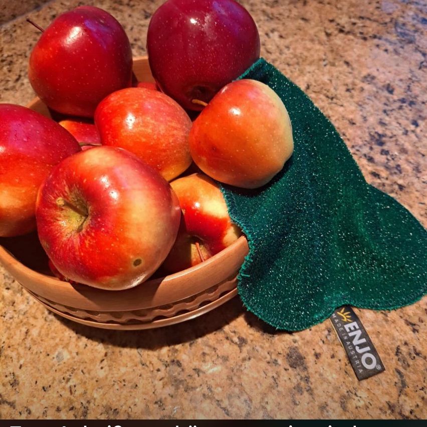 A brown wicker bowl filled with red apples is on a marble countertop. Beside it is a green kitchen cloth with the word 'EnuO' on it.