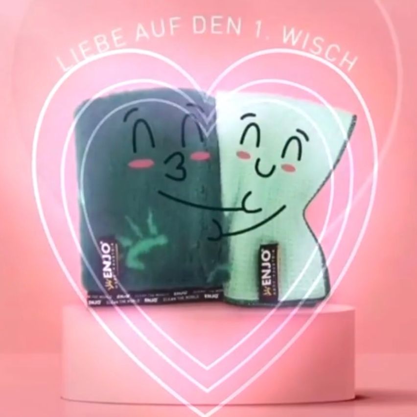 Two ENJO reusable makeup remover pads in green and white with smiling faces inside a heart shape, set on a pink base. The background has the words 'Liebe auf den 1. Wisch'.