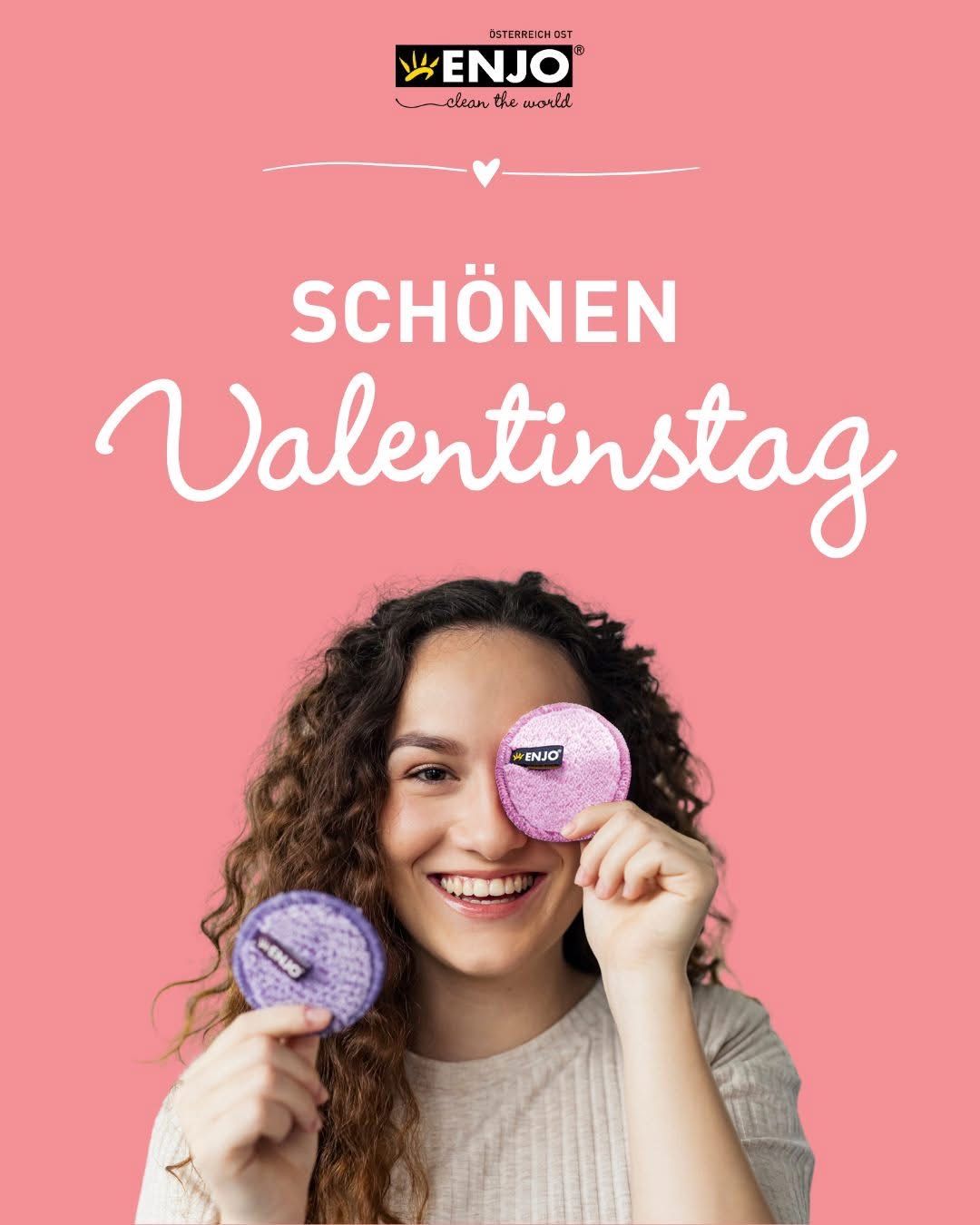 A woman holds two purple facial sponges with the word 'ENJO' on them in front of her face. She is smiling and appears to be advertising a product. The background is pink and features the words 'Schon Valentinstag'.