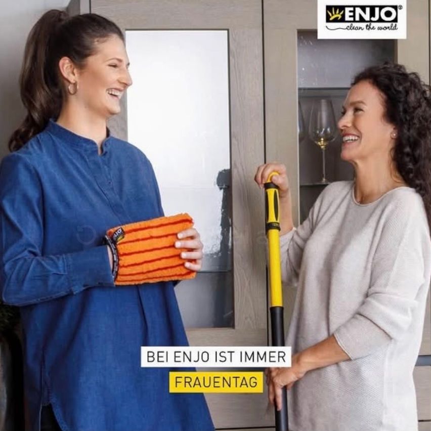 Two women stand in a kitchen. One holds a mop while the other holds a towel. Behind them, a cabinet with a wine glass is visible. The text reads 'At ENJO, it's always Women's Day.'