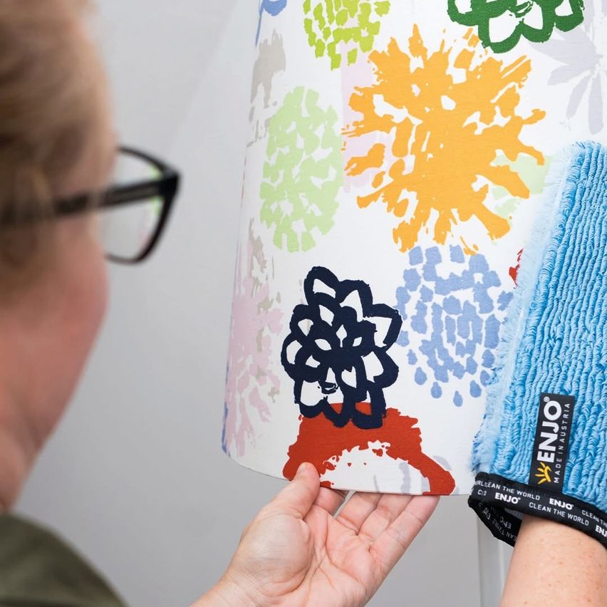Eine Person mit Brille reinigt einen Lampenschirm mit einem blauen Tuch. Der Lampenschirm hat ein farbenfrohes Blumendesign.