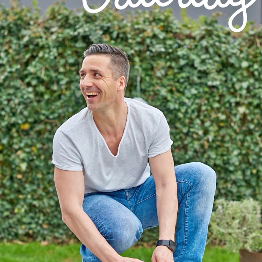 A man wearing a gray t-shirt and blue jeans smiles and squats on a green lawn with a hedge in the background.