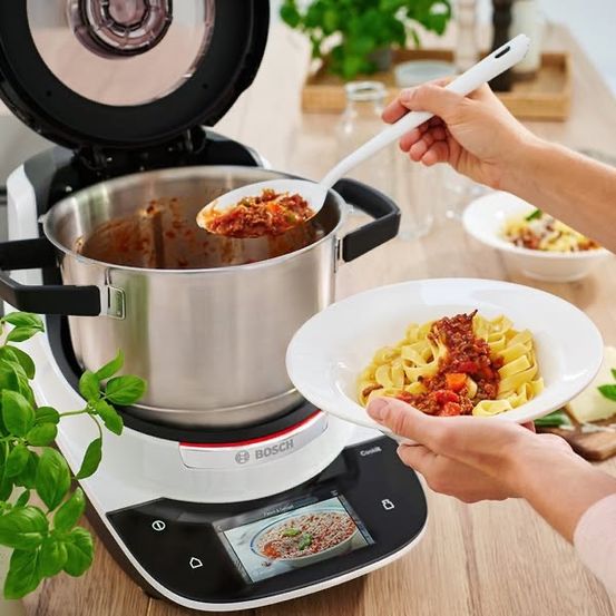 Eine Person hebt Essen aus einem Topf auf einem Bosch Reiskocher in eine Schüssel, mit einer weiteren Schüssel mit Nudeln in der Nähe, alles auf einem Holztisch.