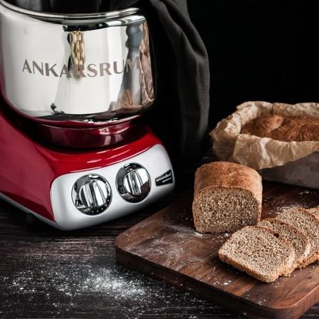 Ein rot-weißes Küchengerät mit der Aufschrift Ankarsrum steht neben einem Holzschneidebrett mit einem Laib Brot darauf.