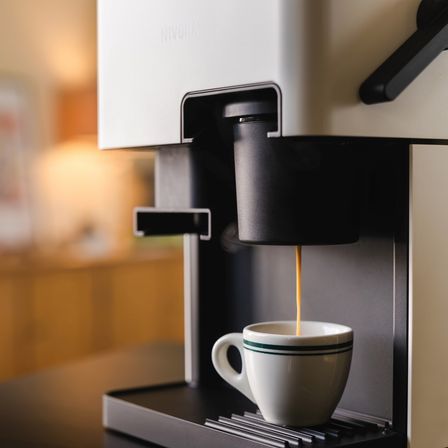 Eine Nivona-Kaffeemaschine brüht Kaffee in eine weiße Tasse mit grünen Streifen. Der Hintergrund ist unscharf und zeigt einen Holzschrank.