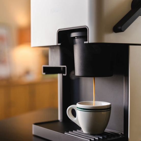 Eine Nivona-Kaffeemaschine brüht Kaffee in eine weiße Tasse mit grünen Streifen. Der Hintergrund ist unscharf und zeigt einen Holzschrank.