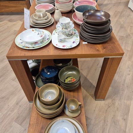 Ein Holztisch mit verschiedenen gestapelten Tellern, Schüsseln und Tassen, einige mit Blumenmustern, auf einem Holzboden.
