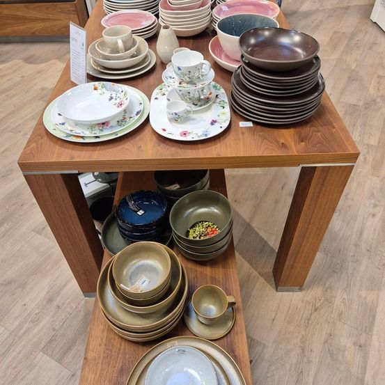 Ein Holztisch mit verschiedenen gestapelten Tellern, Schüsseln und Tassen, einige mit Blumenmustern, auf einem Holzboden.