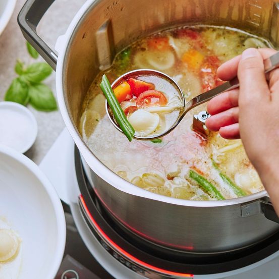 Eine Person benutzt einen Schöpflöffel, um Suppe aus einem Topf auf einem Elektroherd zu schöpfen, gefüllt mit Gemüse wie grüne Bohnen, Tomaten und Basilikum.