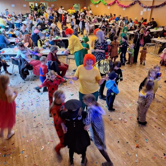 Ein großer Innenraum voller Menschen, viele in Kostümen, tanzen und werfen Konfetti. Tische mit Essen und Getränken umgeben den Rand. Eine Frau in einem gelben Shirt lächelt in die Kamera.