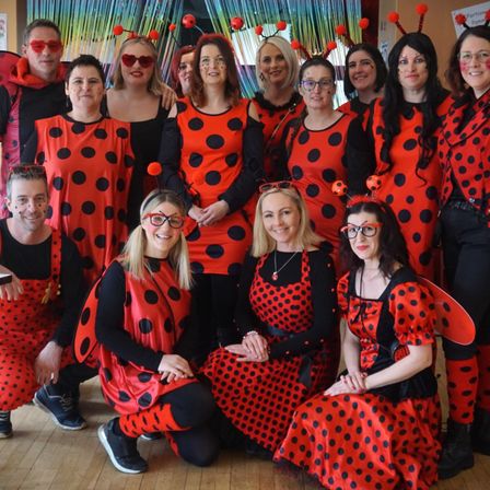 Eine Gruppe von Erwachsenen in roten und schwarzen Tupfen-Marienkäferkostümen mit Flügeln, die für ein Foto posieren. Der Hintergrund hat einen bunten Vorhang.