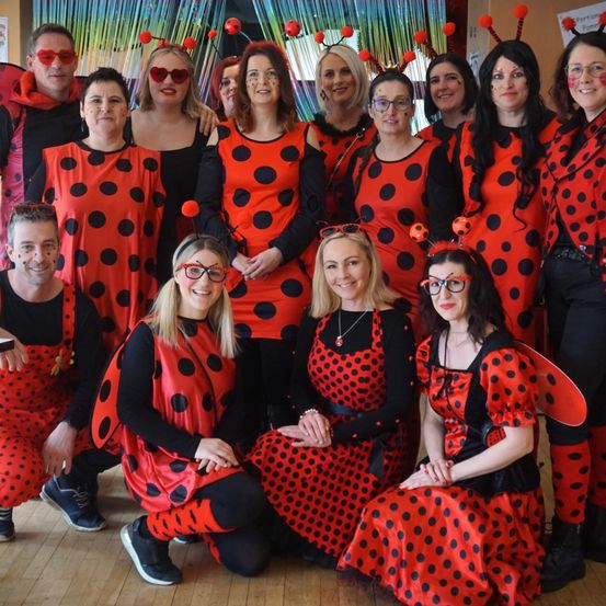 Eine Gruppe von Erwachsenen in roten und schwarzen Tupfen-Marienkäferkostümen mit Flügeln, die für ein Foto posieren. Der Hintergrund hat einen bunten Vorhang.
