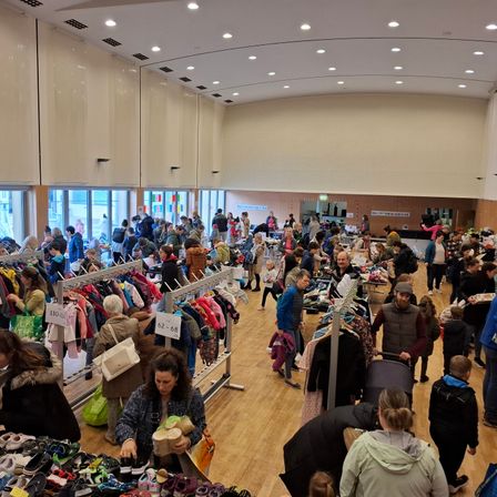 Ein großer Innenbereich voller Menschen, die Kleidung und Schuhe kaufen. Viele Artikel hängen an Kleiderbügeln und Tischen, mit einer Mischung aus Winterkleidung und Schuhen. Der Boden ist aus Holz und die Wände sind mit Glasfenstern geschmückt.