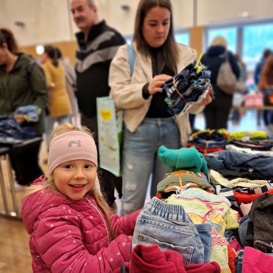 Ein kleines Mädchen in einer rosa Jacke lächelt, während es auf einem Tisch voller Kleidung bei einer Indoor-Veranstaltung stöbert. Hinter ihr hält eine Frau einen Haufen Kleidung, und verschiedene Menschen gehen umher.
