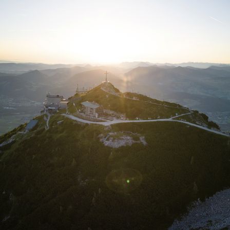 Ein Luftbild eines Berggipfels mit einem Kreuz auf der Spitze, einem kleinen Gebäude und einem Weg, der dorthin führt. Die Sonne geht im Hintergrund unter.