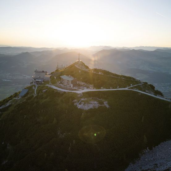 An aerial view of a mountain peak with a cross at the top, a small building, and a road leading to it. The sun is setting in the background.