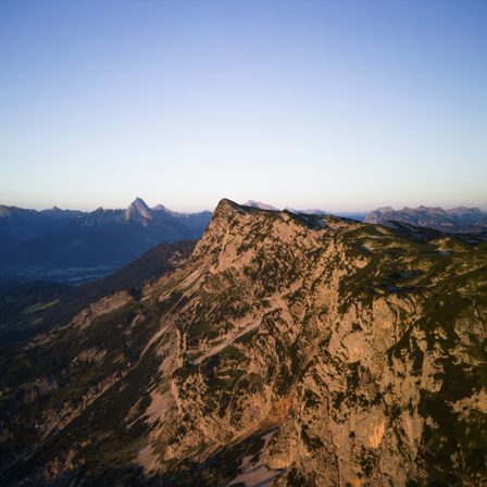 Luftaufnahme einer Berglandschaft mit steilen Klippen, rauem Gelände und spärlicher Vegetation unter klarem Himmel.