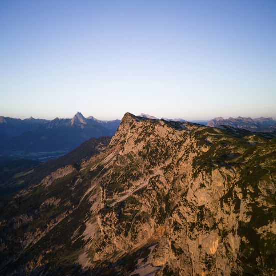 Aerial view of a mountainous landscape with steep cliffs, rugged terrain, and sparse vegetation under a clear sky.